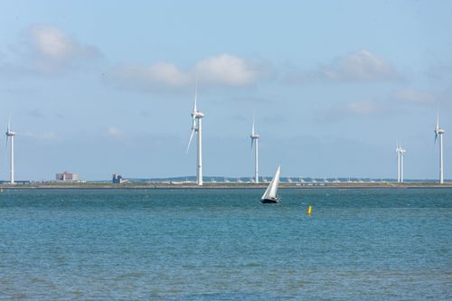 Free A sailboat glides near offshore wind turbines under a bright sky, highlighting renewable energy in a coastal setting. Stock Photo