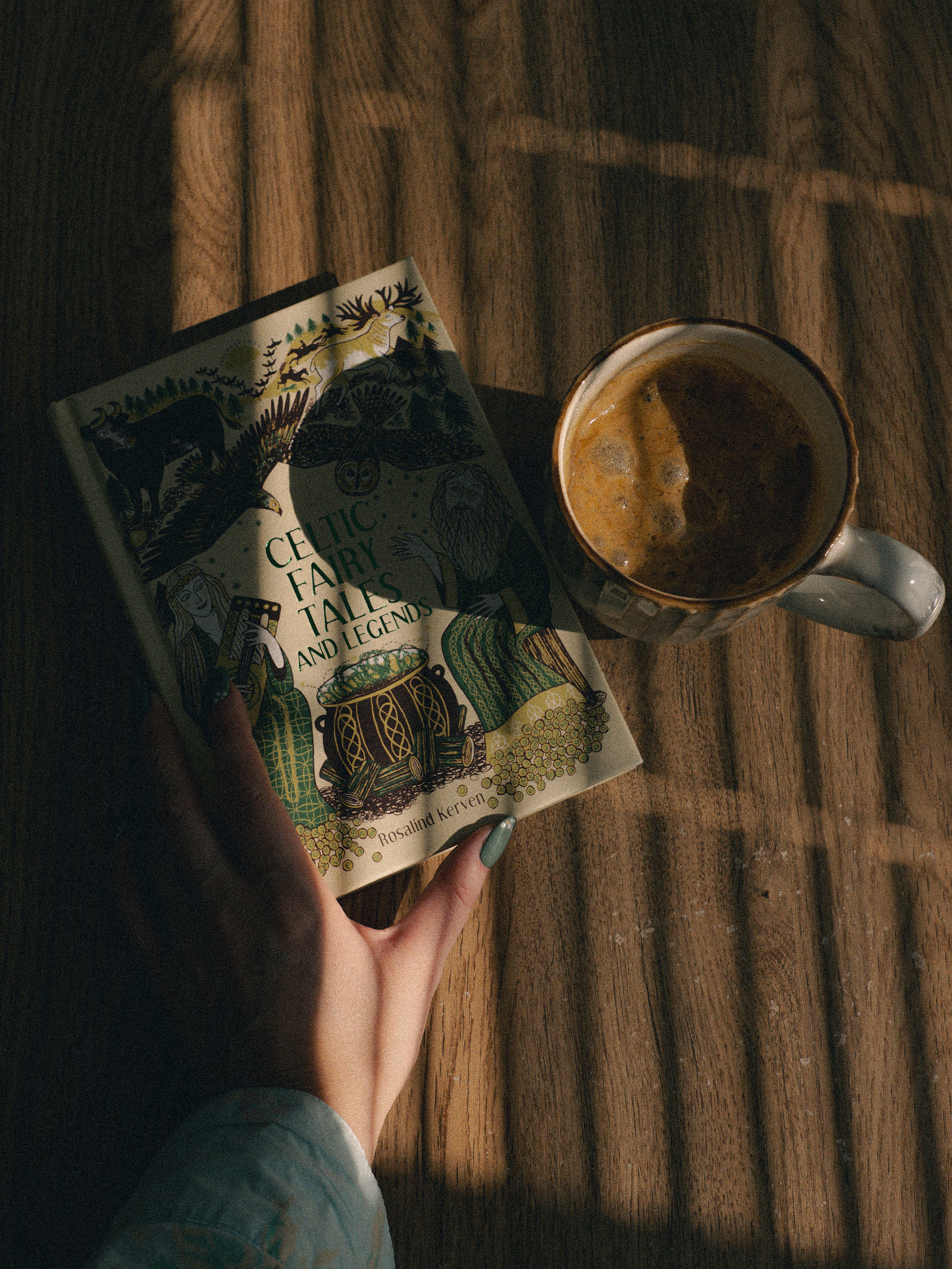 A person holding a book titled 'Celtic Fairy Tales and Legends' next to a coffee cup on a wooden table.