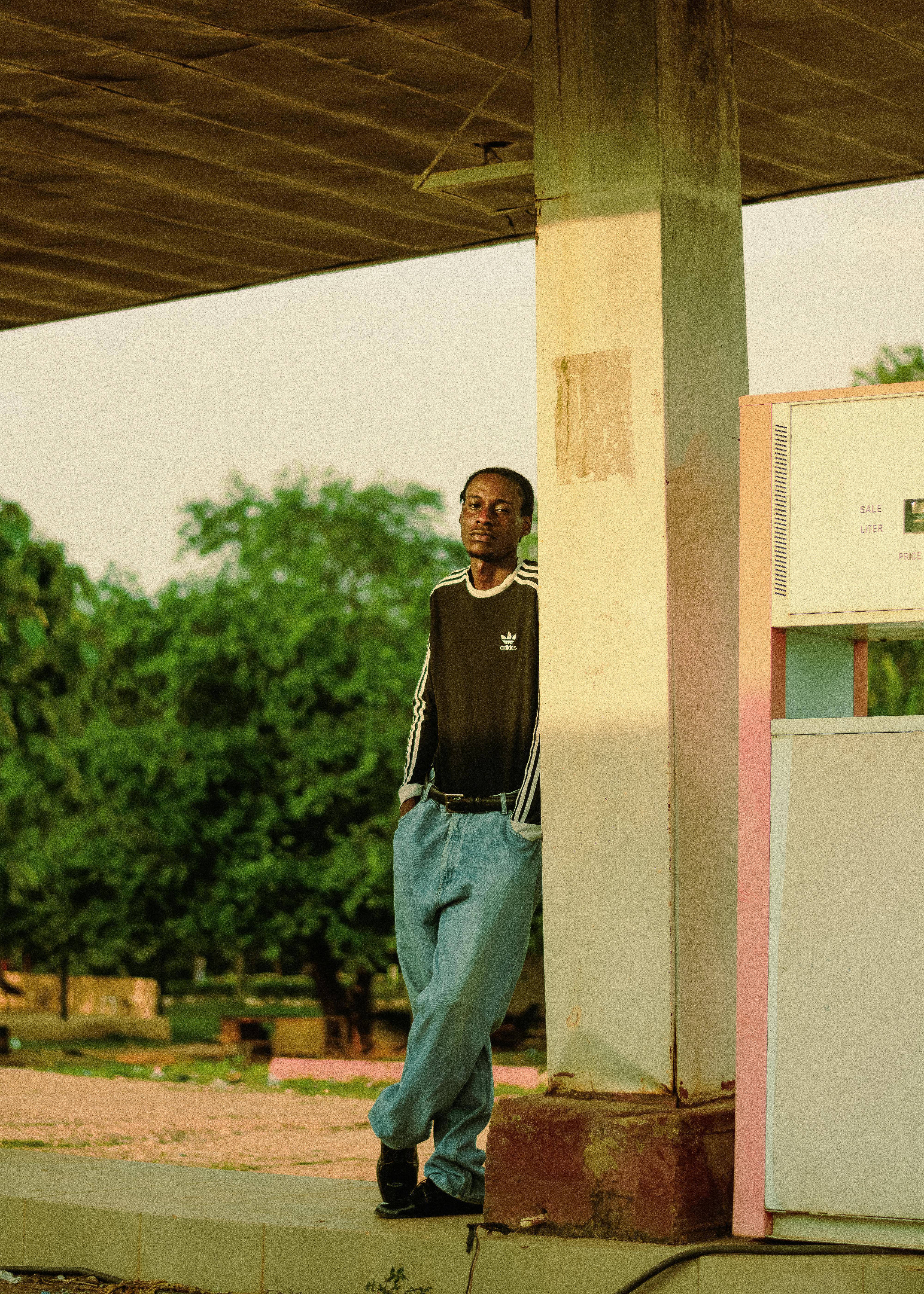 Young man in casual attire leaning against a gas station pillar in a tranquil outdoor setting.