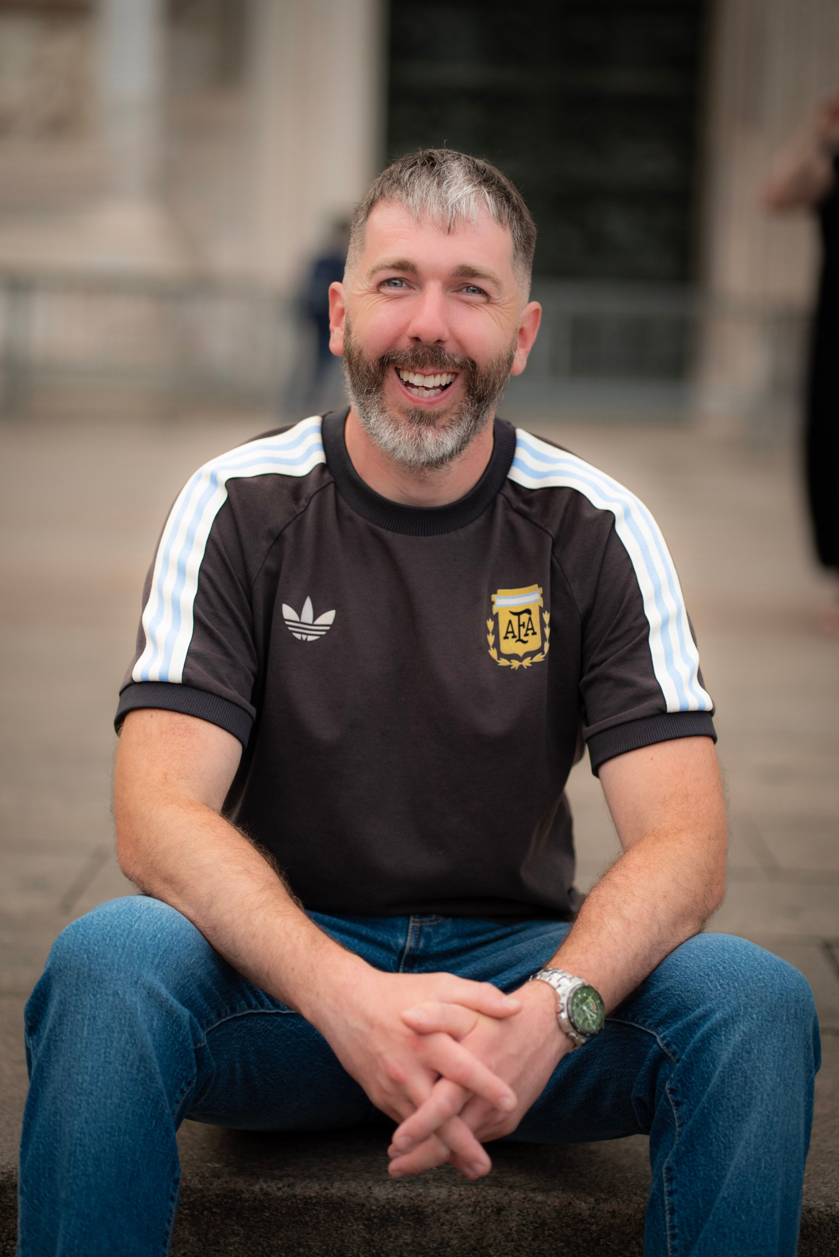 A cheerful adult man sitting outdoors in Milan, Italy, wearing a casual sports shirt.