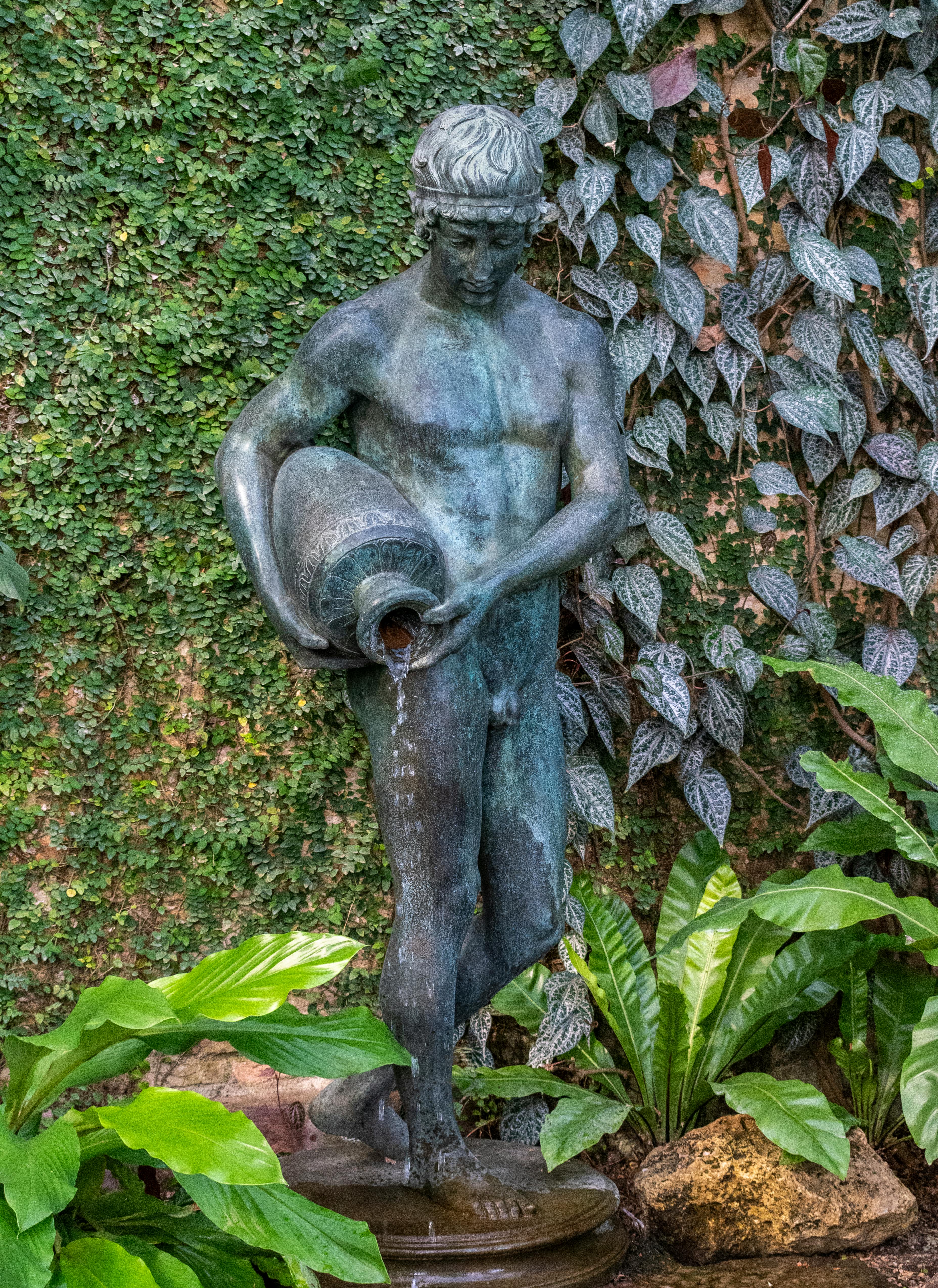 Free Elegant bronze sculpture of a man pouring water in a verdant garden setting, creating a classic aesthetic. Stock Photo