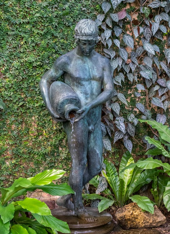 Free Elegant bronze sculpture of a man pouring water in a verdant garden setting, creating a classic aesthetic. Stock Photo