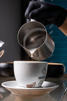 Barista using a pitcher to pour creamy steamed milk into a white coffee cup, creating latte art.