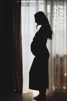 Silhouette of a pregnant woman standing by a window, showcasing maternity.