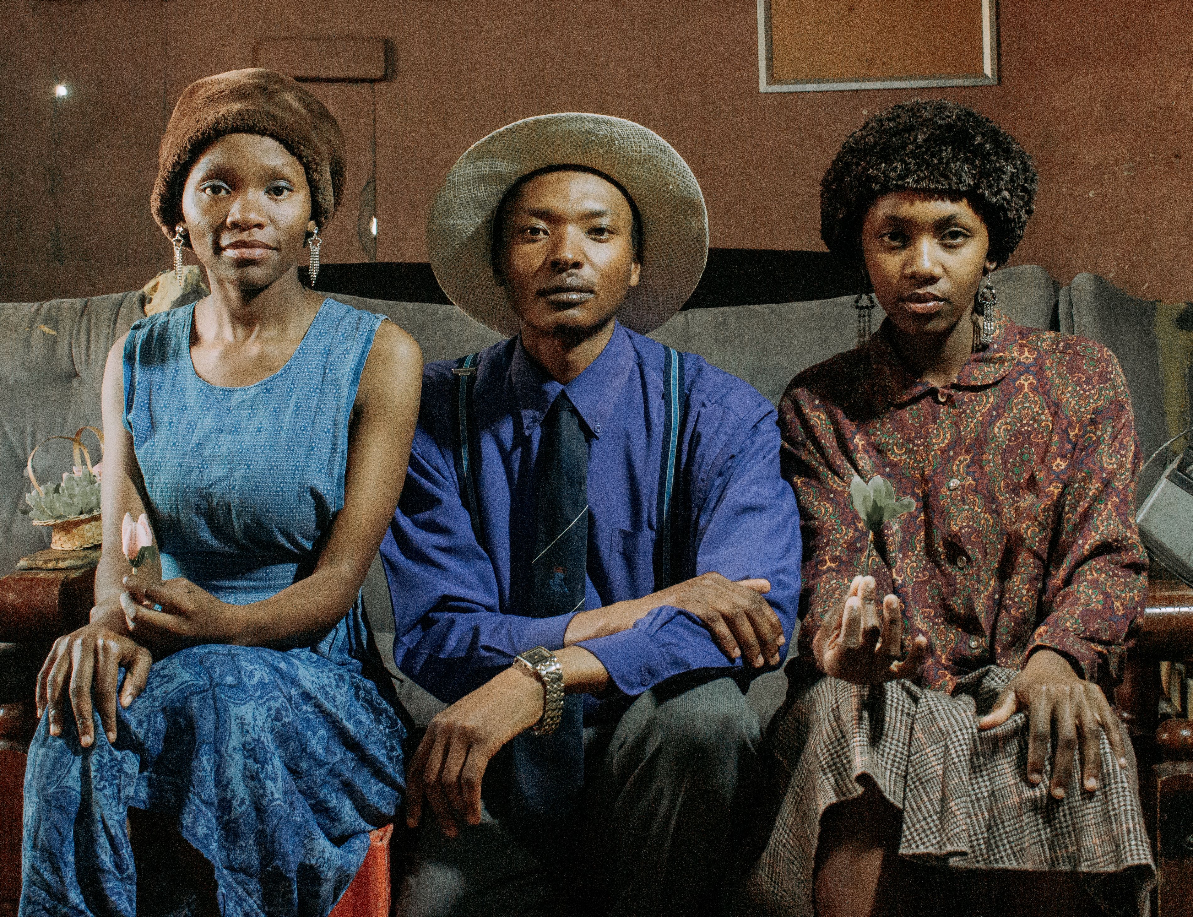 Three individuals dressed in vintage clothing pose indoors with succulents, showcasing retro style.