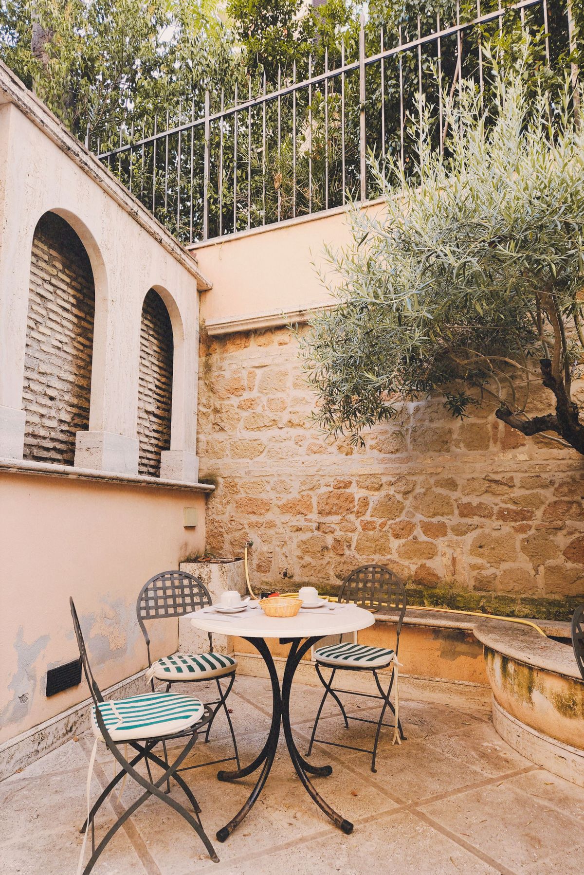 Sunny terrace with an iron table, chairs and an olive tree beside a stone wall