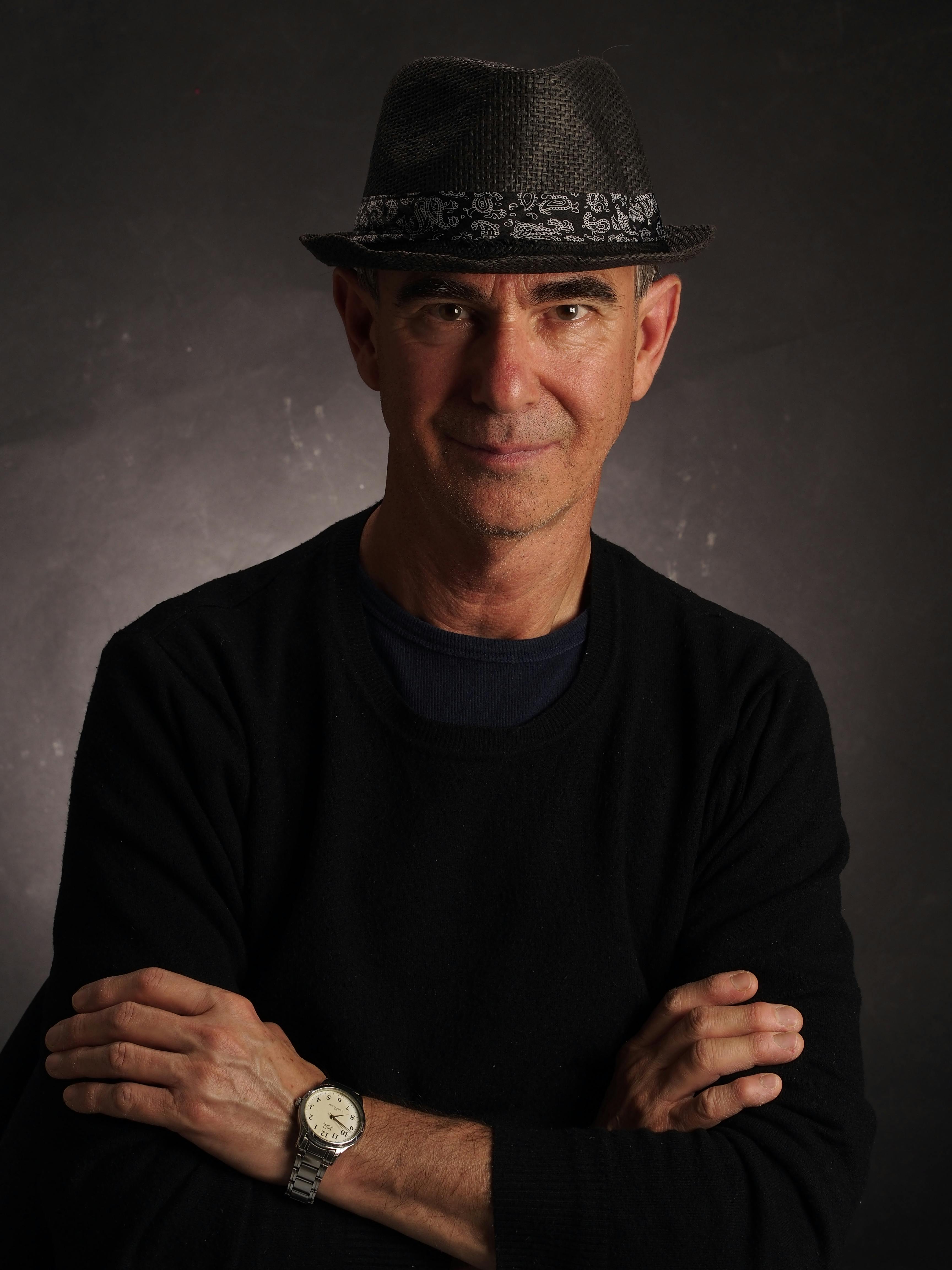 Confident man in a dark fedora, crossing arms, against a moody backdrop.