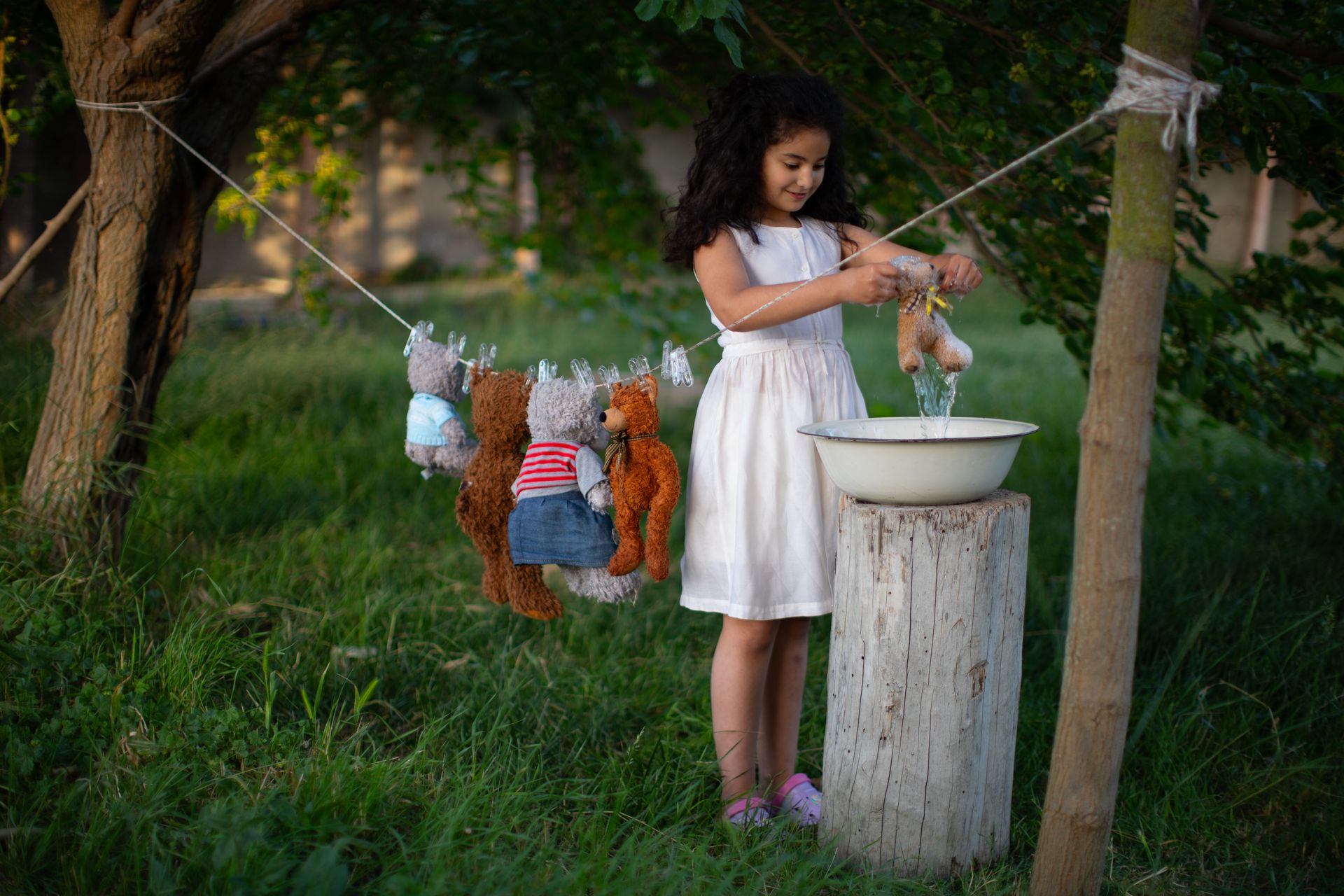 A young girl in a white dress washes teddy bears outdoors on a sunny summer day.