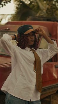 Stylish young woman in white shirt and tie poses confidently by a red vintage car.
