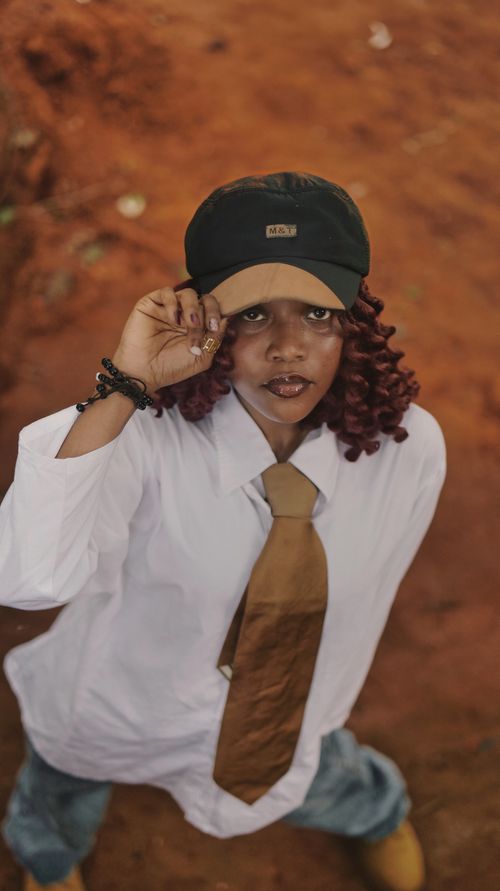 Free Fashionable individual in casual attire with tie and cap, standing on red soil outdoor background. Stock Photo