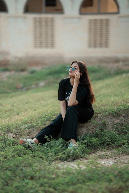 Free Portrait of a young woman sitting thoughtfully on grass outdoors, wearing casual attire and sunglasses. Stock Photo