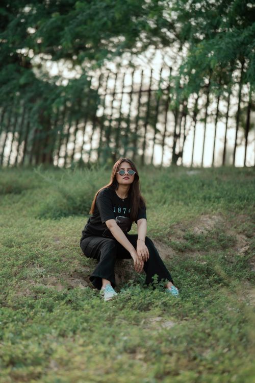 Free Casual young woman seated on grass in a park, wearing sunglasses and relaxed clothing. Stock Photo