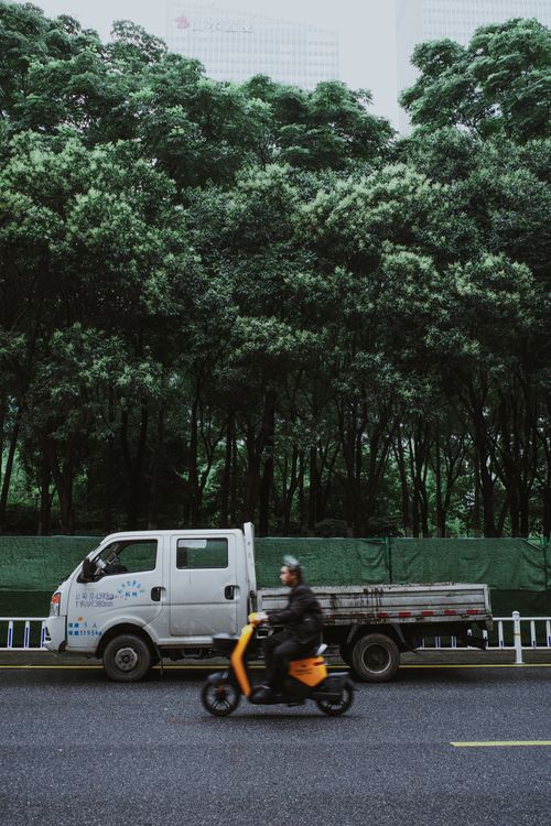 Free Dynamic urban scene featuring a moving scooter and parked truck against a lush green backdrop. Stock Photo
