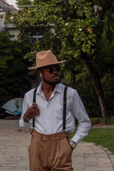 Man wearing a hat and sunglasses with a vintage style outfit in an outdoor setting.