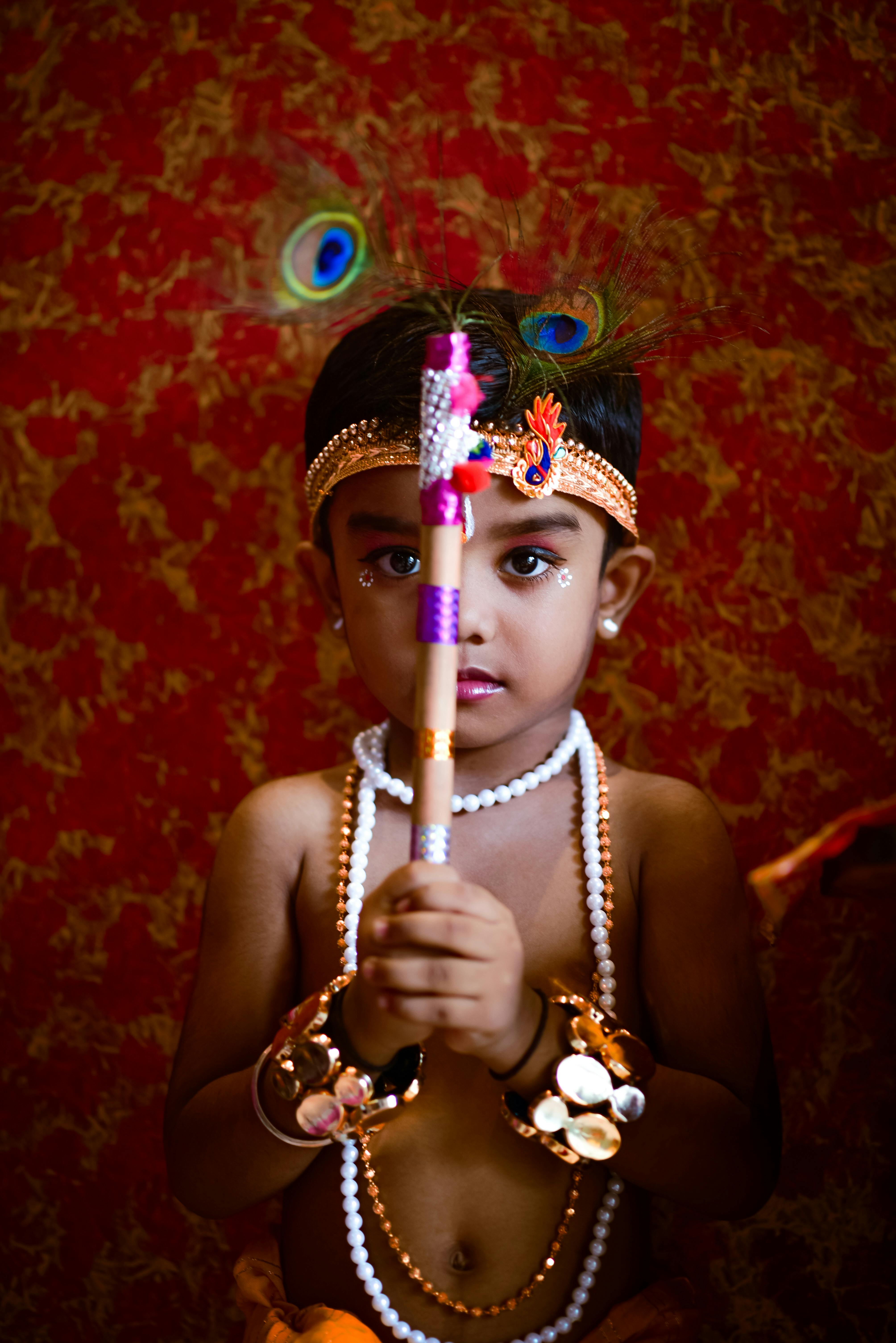 A child dressed as Lord Krishna holding a flute, depicting a traditional Hindu festival scene.