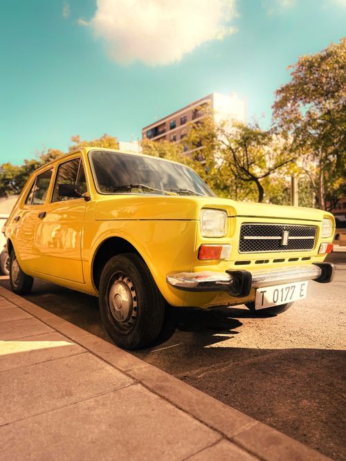 Free Yellow vintage car parked on a sunny street with trees and buildings in the background. Stock Photo