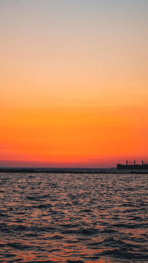 Free Vibrant orange sunset over calm ocean waves with distant cargo ship silhouette. Stock Photo