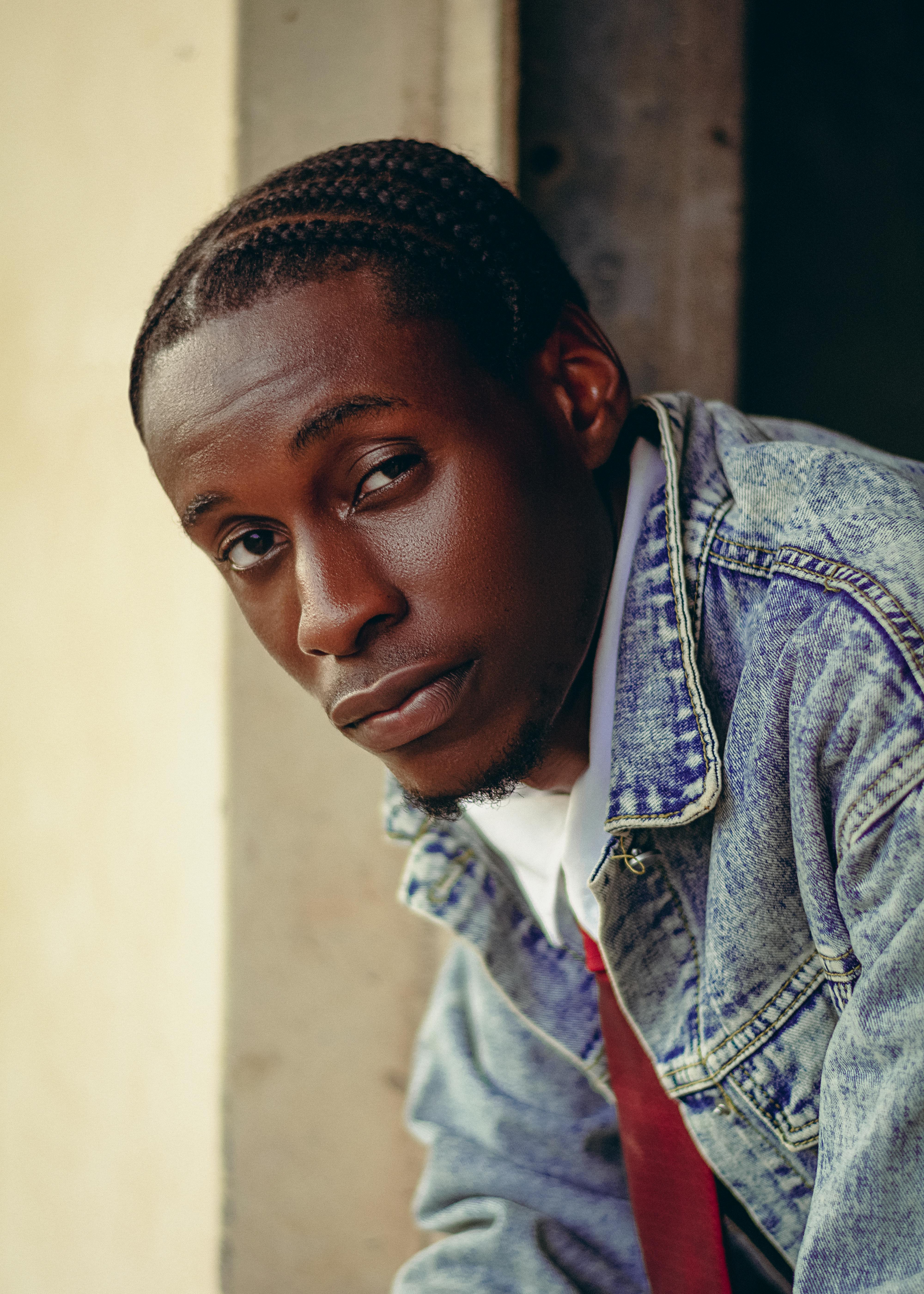 Portrait of a young man in a denim jacket with braided hair, gazing thoughtfully.