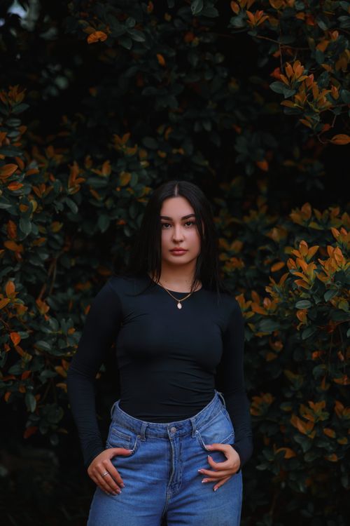 Free A young woman in denim jeans and black top stands confidently against a lush green background. Stock Photo