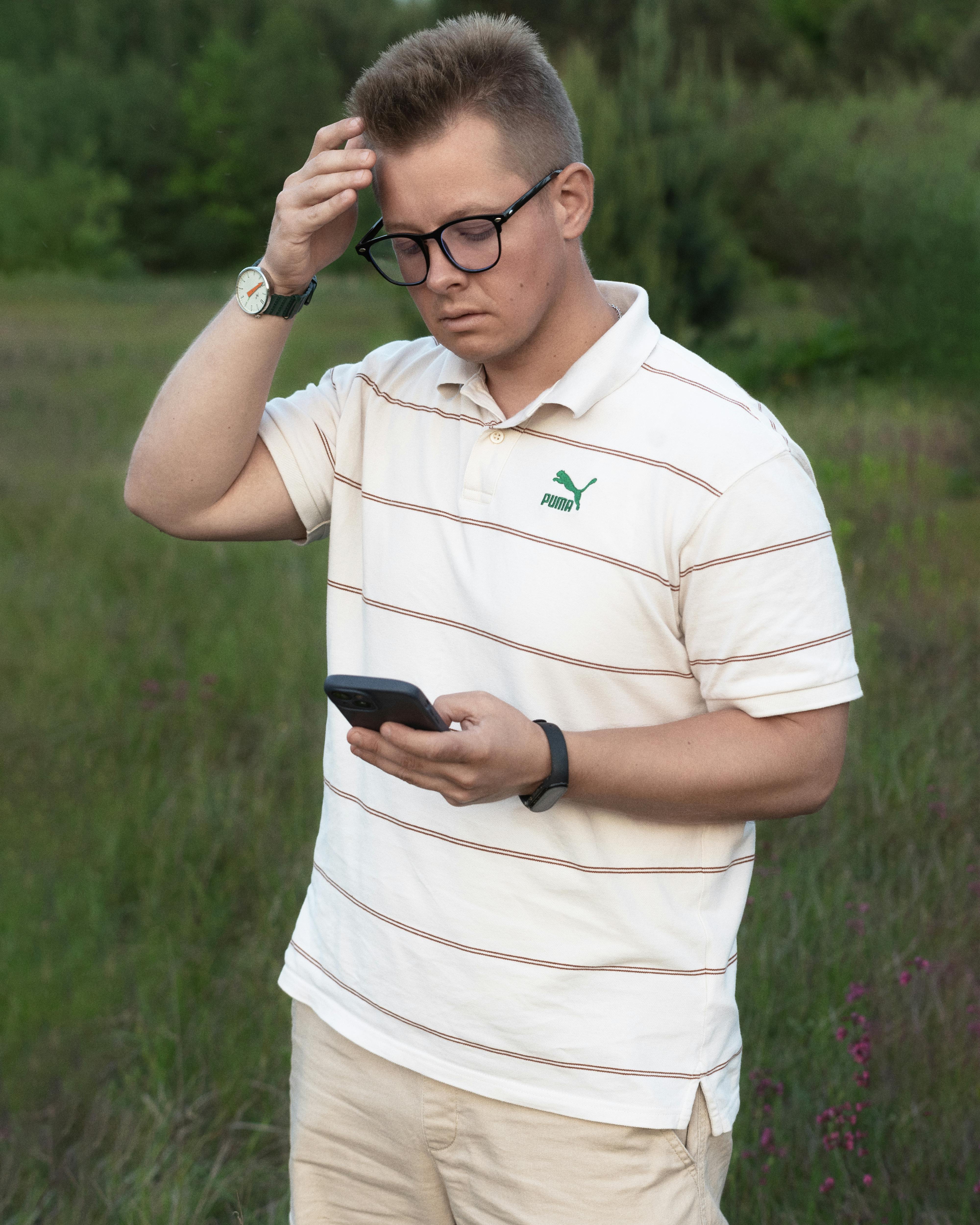A young man in casual wear checking his smartphone in a natural outdoor setting, looking thoughtful.