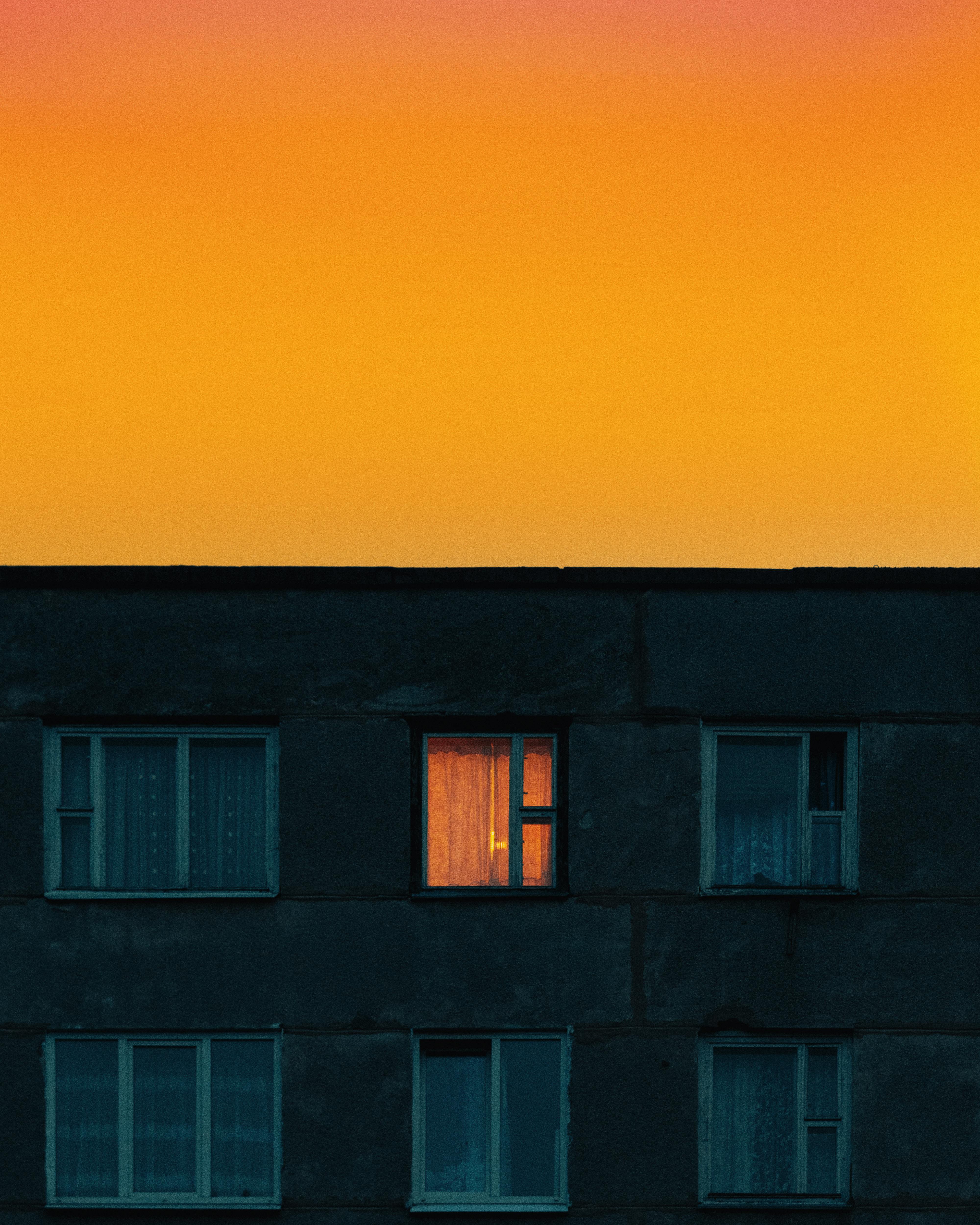 A vibrant orange sunset over a dark building facade with a single illuminated window.