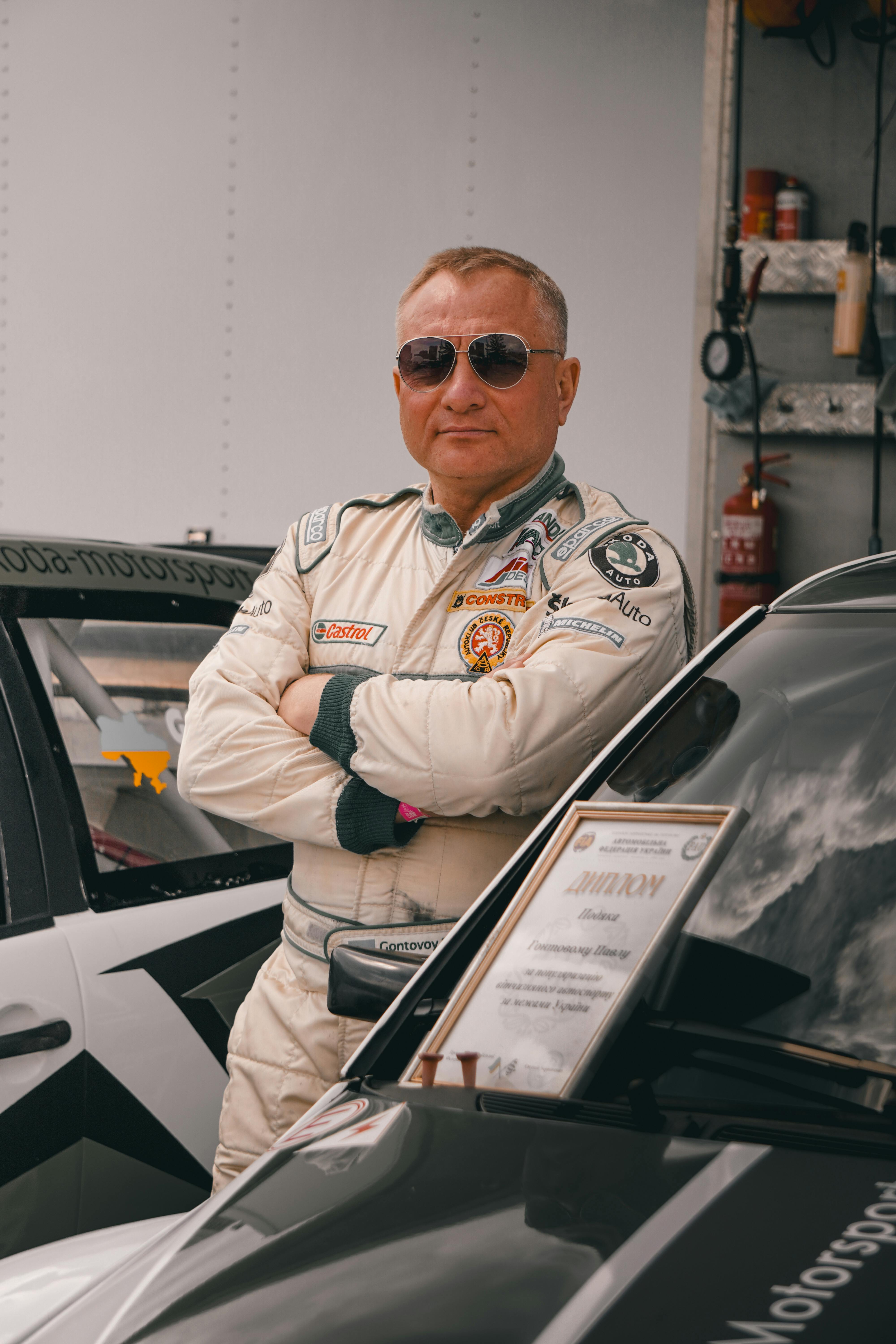 Confident race car driver wearing racing suit and sunglasses poses next to a car and trophy at an event in Kyiv.