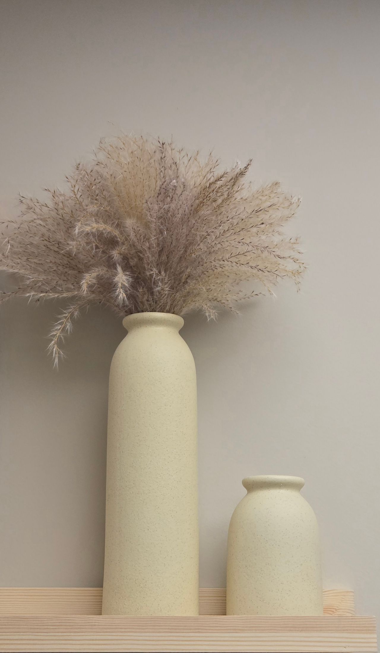 Feathery dried grass bouquet in a tall cream vase on a wooden shelf beside a smaller vase