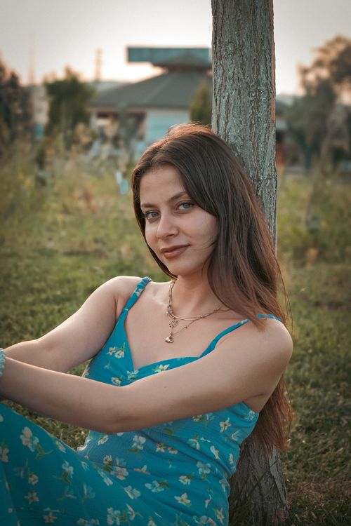 Free Portrait of a young woman sitting against a tree in a summer dress, enjoying a peaceful day outdoors. Stock Photo
