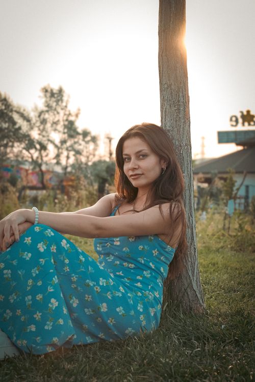 Free Woman in floral dress relaxing by a tree during sunset creating a serene summer vibe. Stock Photo