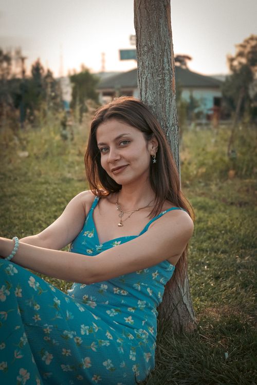 Free Smiling young woman in blue dress sitting by a tree outdoors during sunset. Stock Photo
