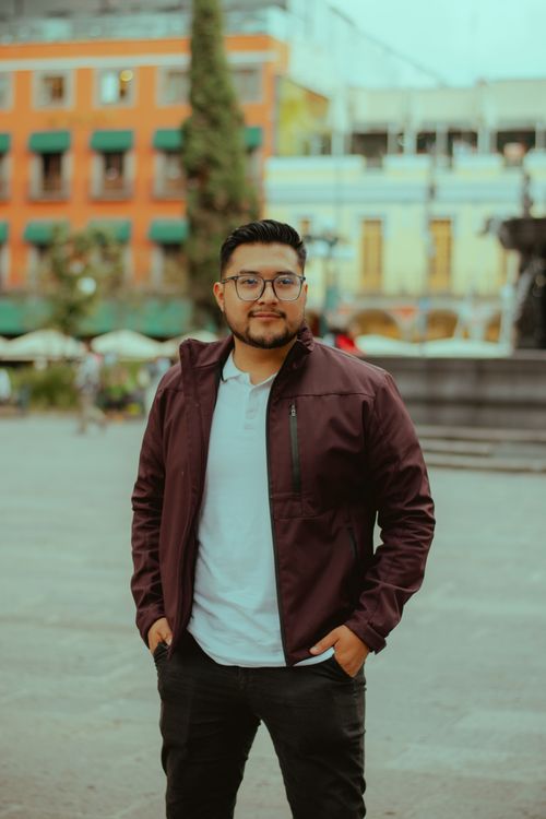 Free Young man in a purple jacket posing confidently in a city square, featuring colorful buildings. Stock Photo