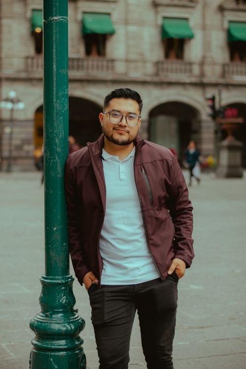 Free Portrait of a stylish young man casually leaning against a street lamp in an urban environment. Stock Photo