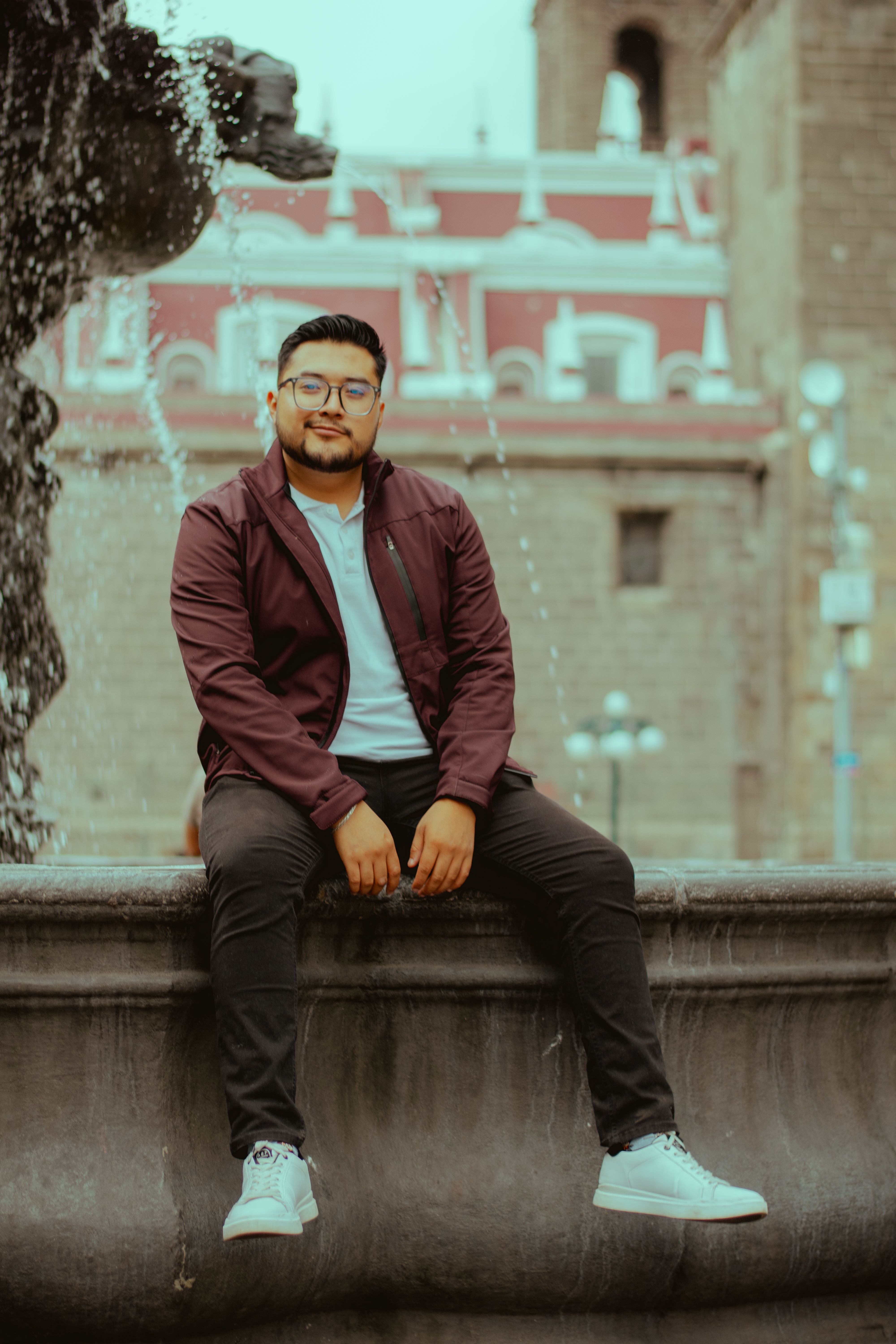 Young man in casual attire sitting by decorative fountain with historic architecture backdrop.