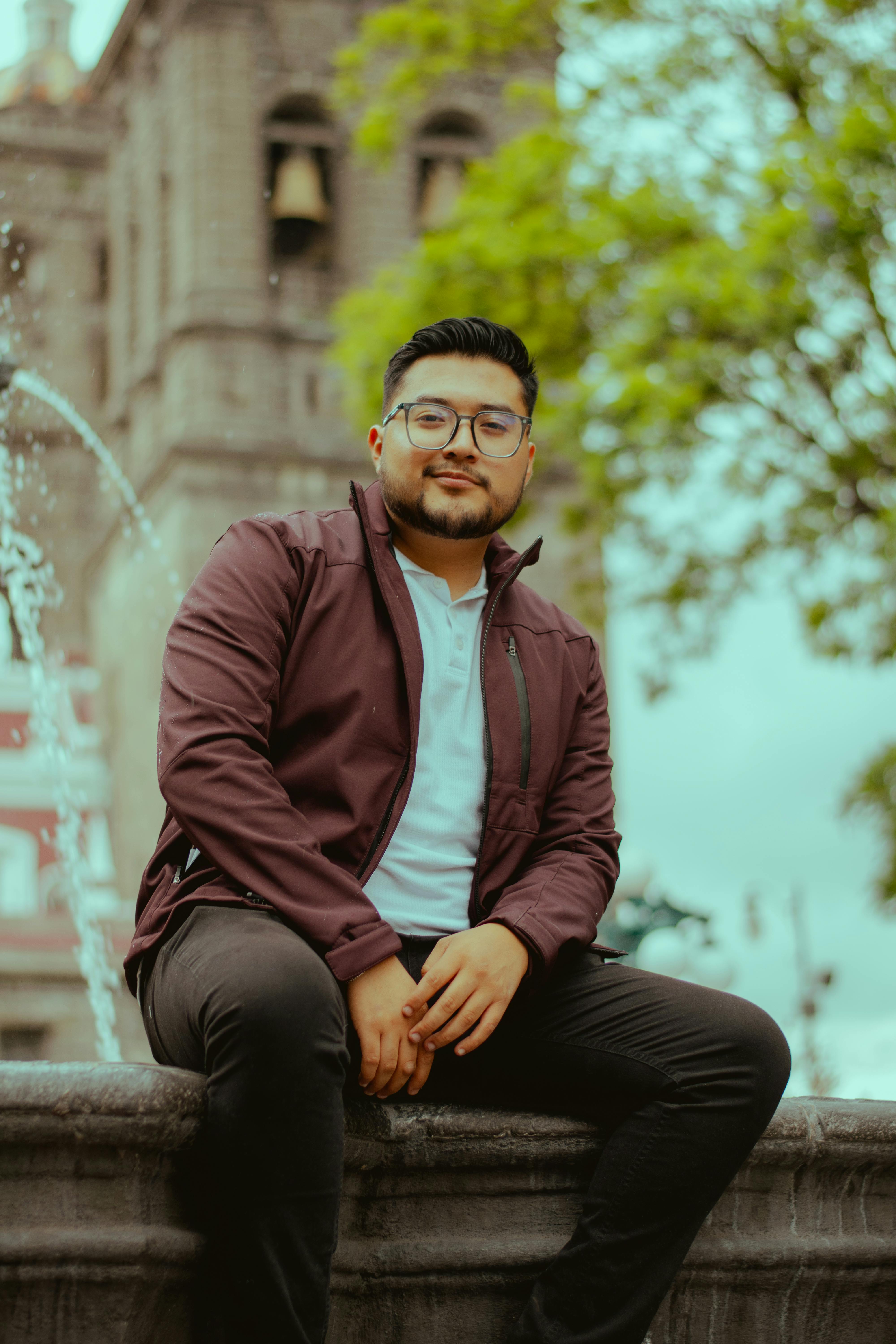 Young man sitting by a fountain with a historic building in the background, outdoors.
