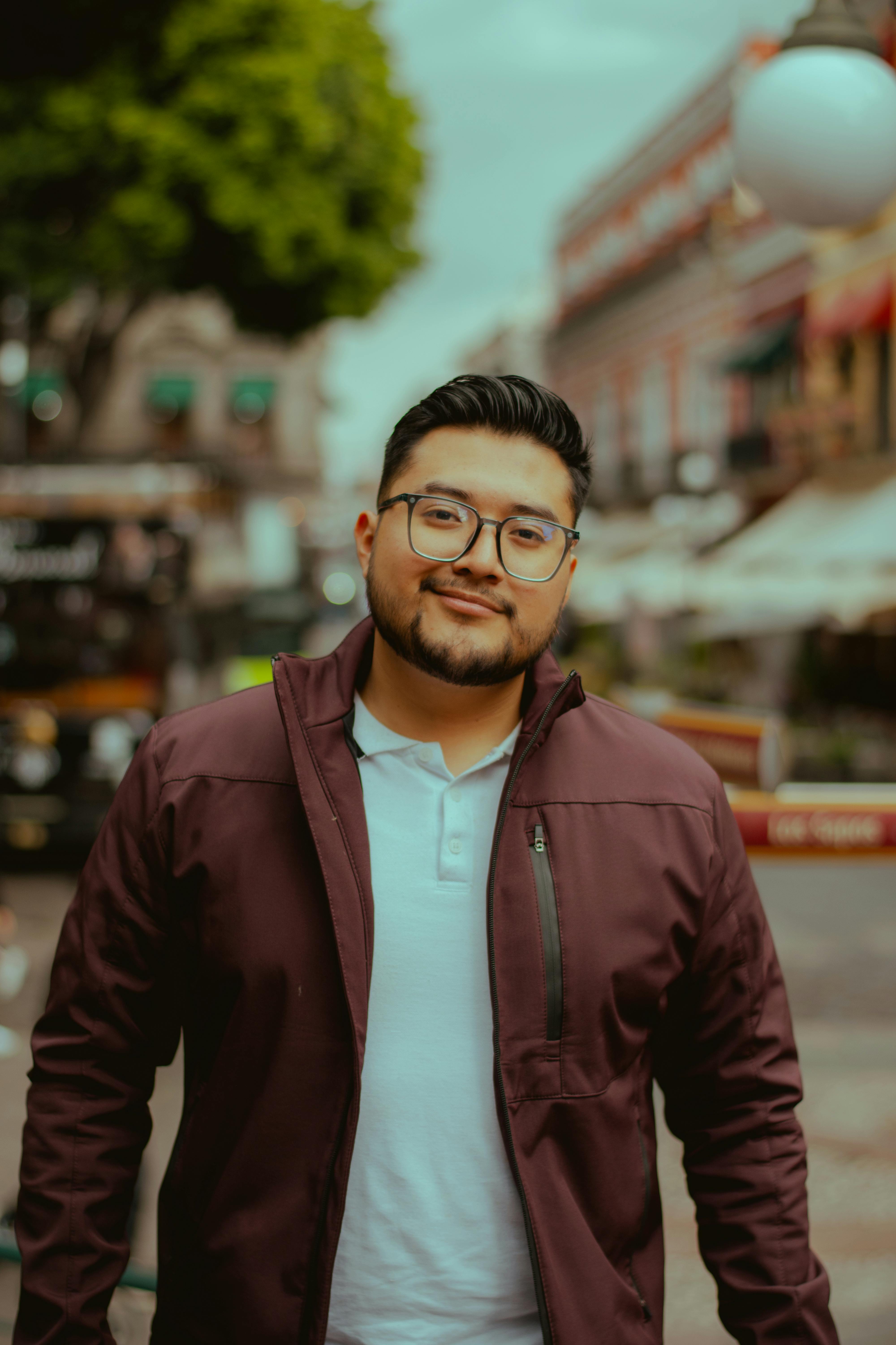 De franc Retrat informal d'un home amb ulleres i jaqueta grana dempeus en un carrer urbà bulliciós. Foto d'estoc