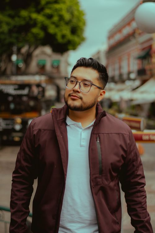 Free Young man in glasses and jacket walking in a lively city street with blurred background. Stock Photo