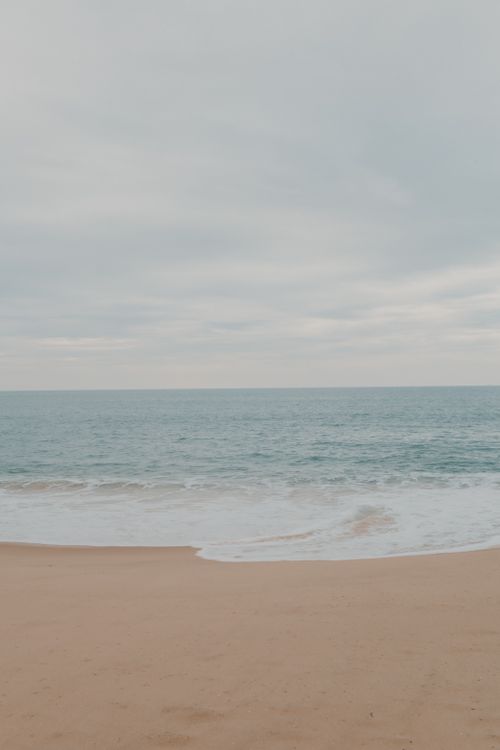 Free A serene beach view featuring gentle waves lapping against a sandy shore under a cloudy sky. Stock Photo