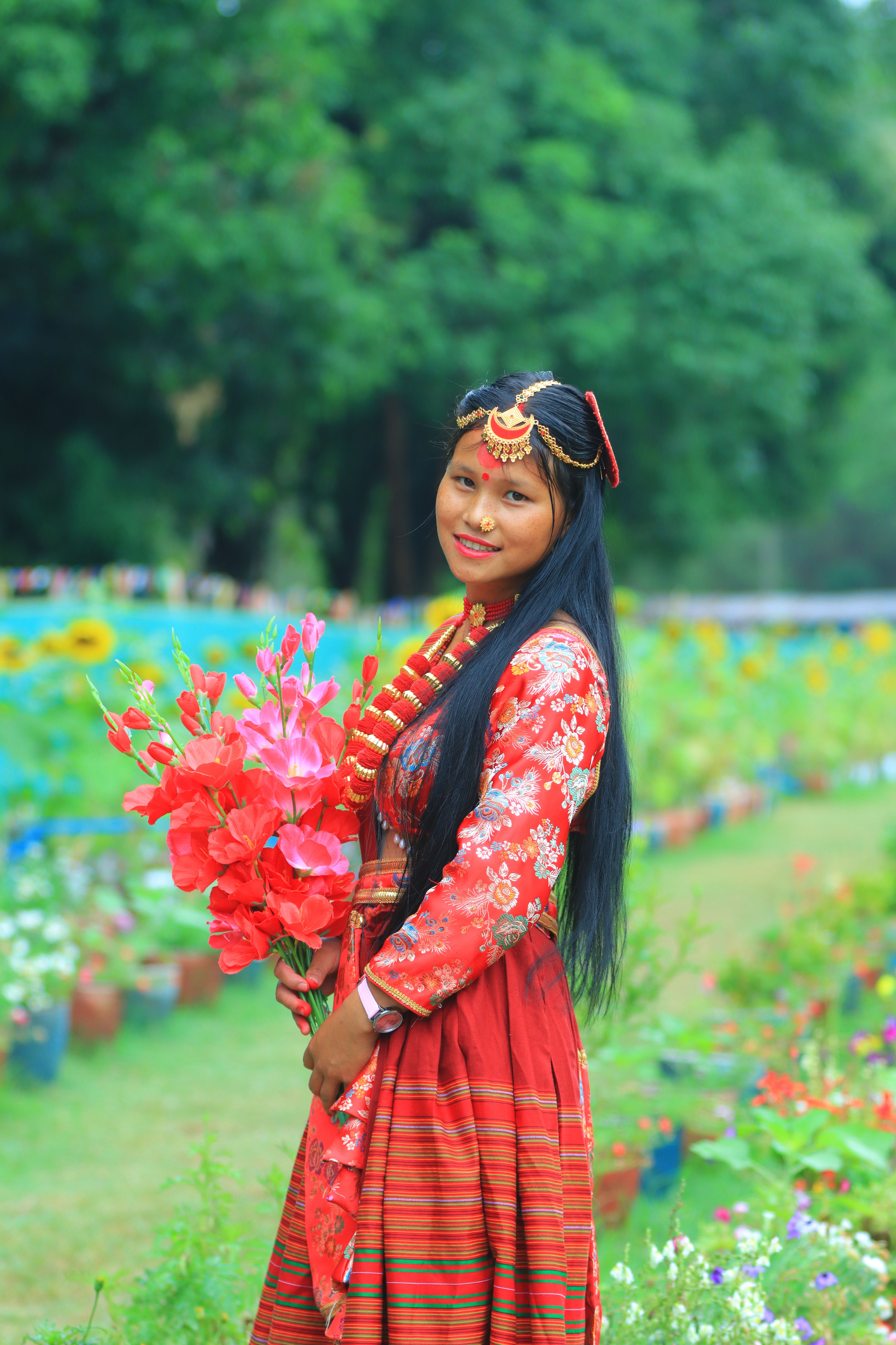 A woman in cultural attire holds vibrant flowers in a lush garden setting, exuding joy.