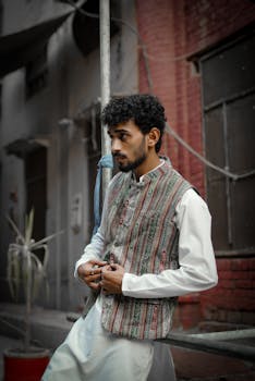 Stylish man in traditional attire poses thoughtfully against urban backdrop, highlighting cultural fashion.