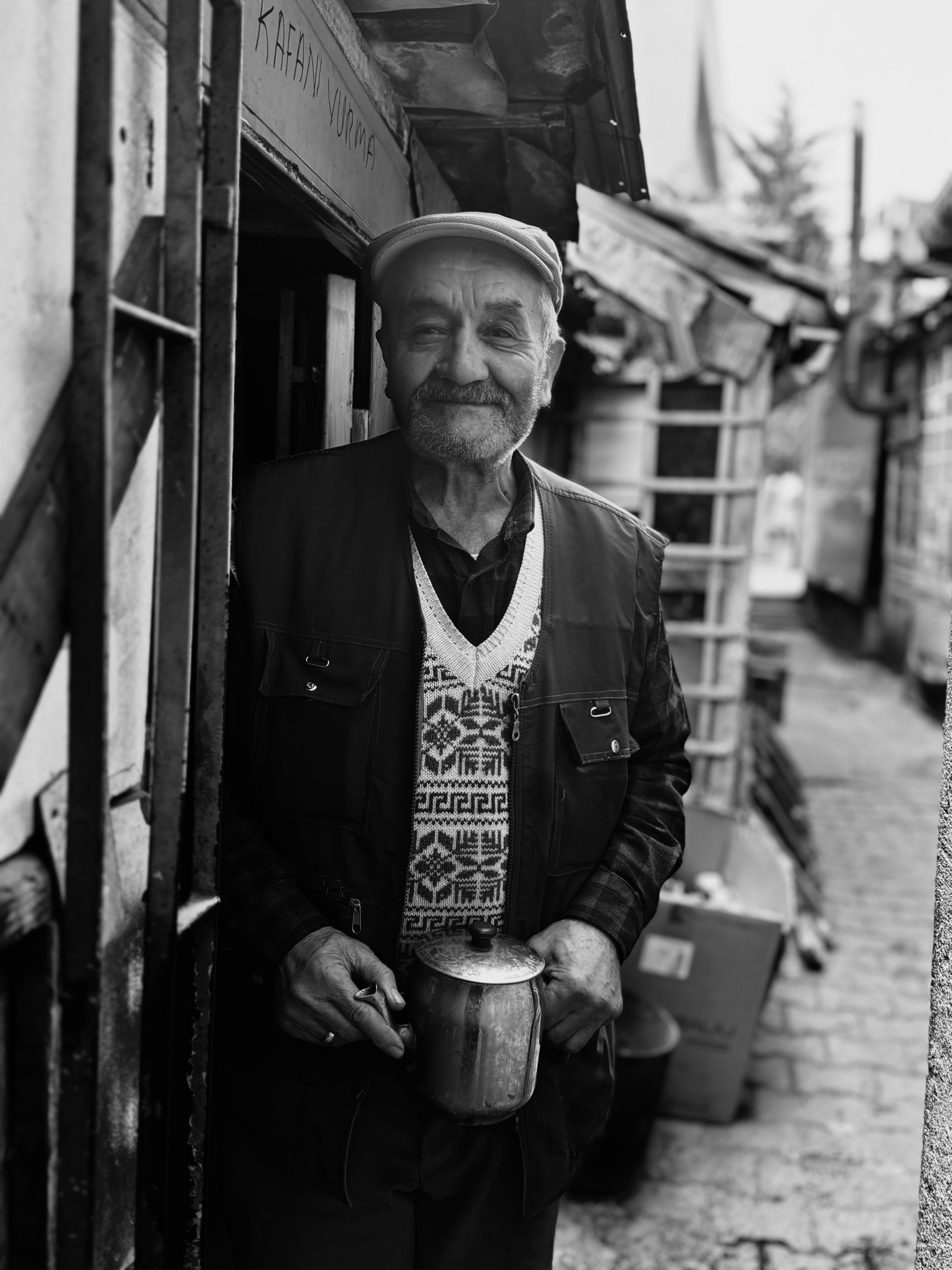 Black and white portrait of an elderly man holding a mug in a rustic alleyway.