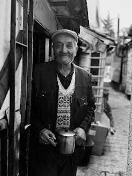 Black and white portrait of an elderly man holding a mug in a rustic alleyway.