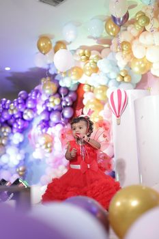Adorable toddler in red dress at a vibrant birthday party, surrounded by balloons.