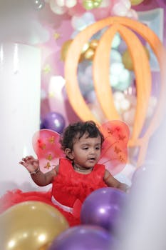 Charming baby in red dress with butterfly wings amidst colorful balloons at a vibrant birthday party.