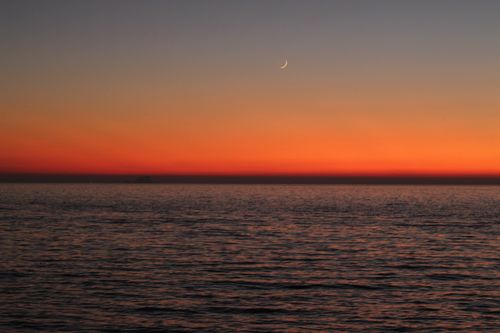 Free Beautiful orange sunset over calm ocean with crescent moon in sky. Stock Photo