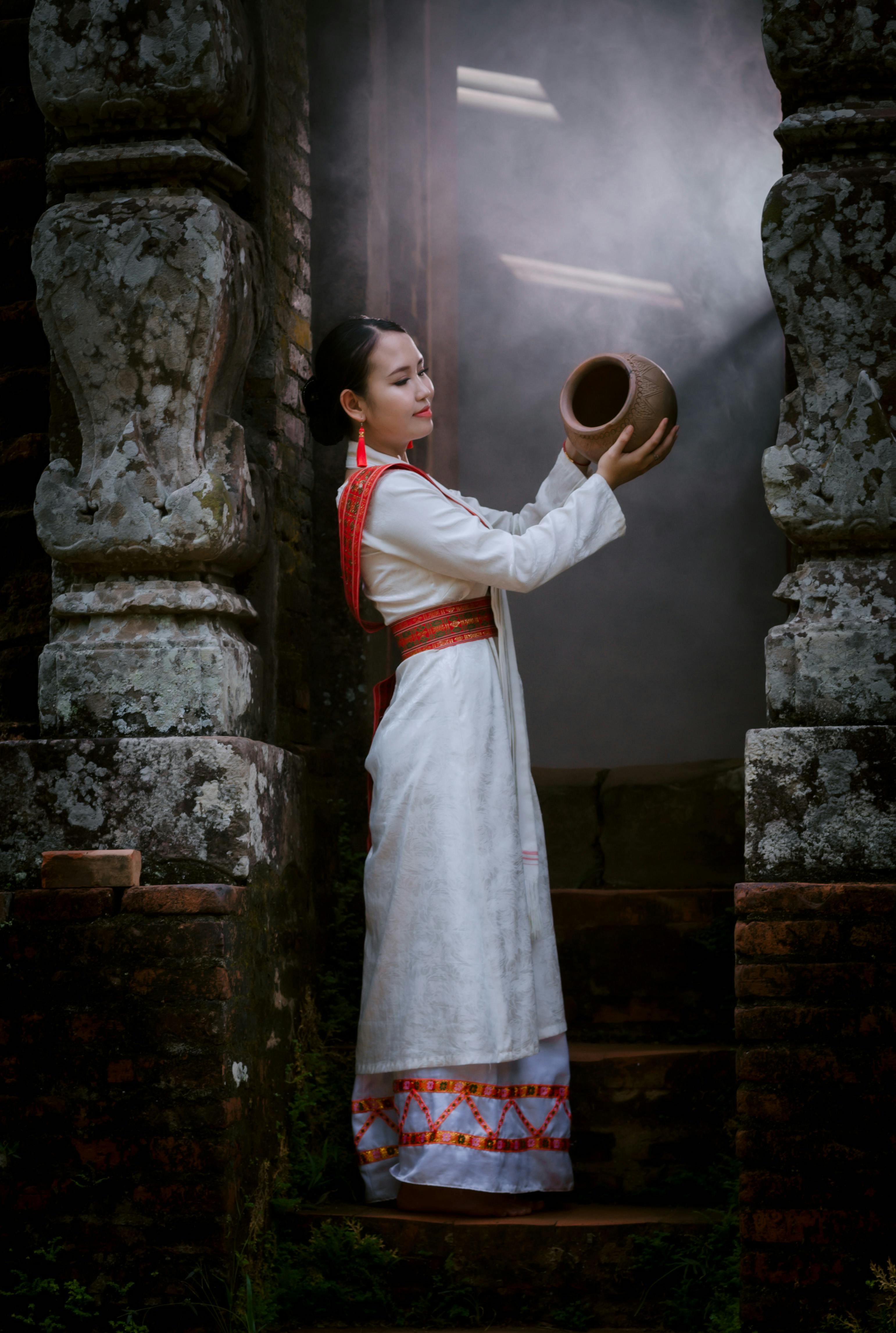 A woman in traditional ethnic attire stands holding a clay pot in a rustic setting.