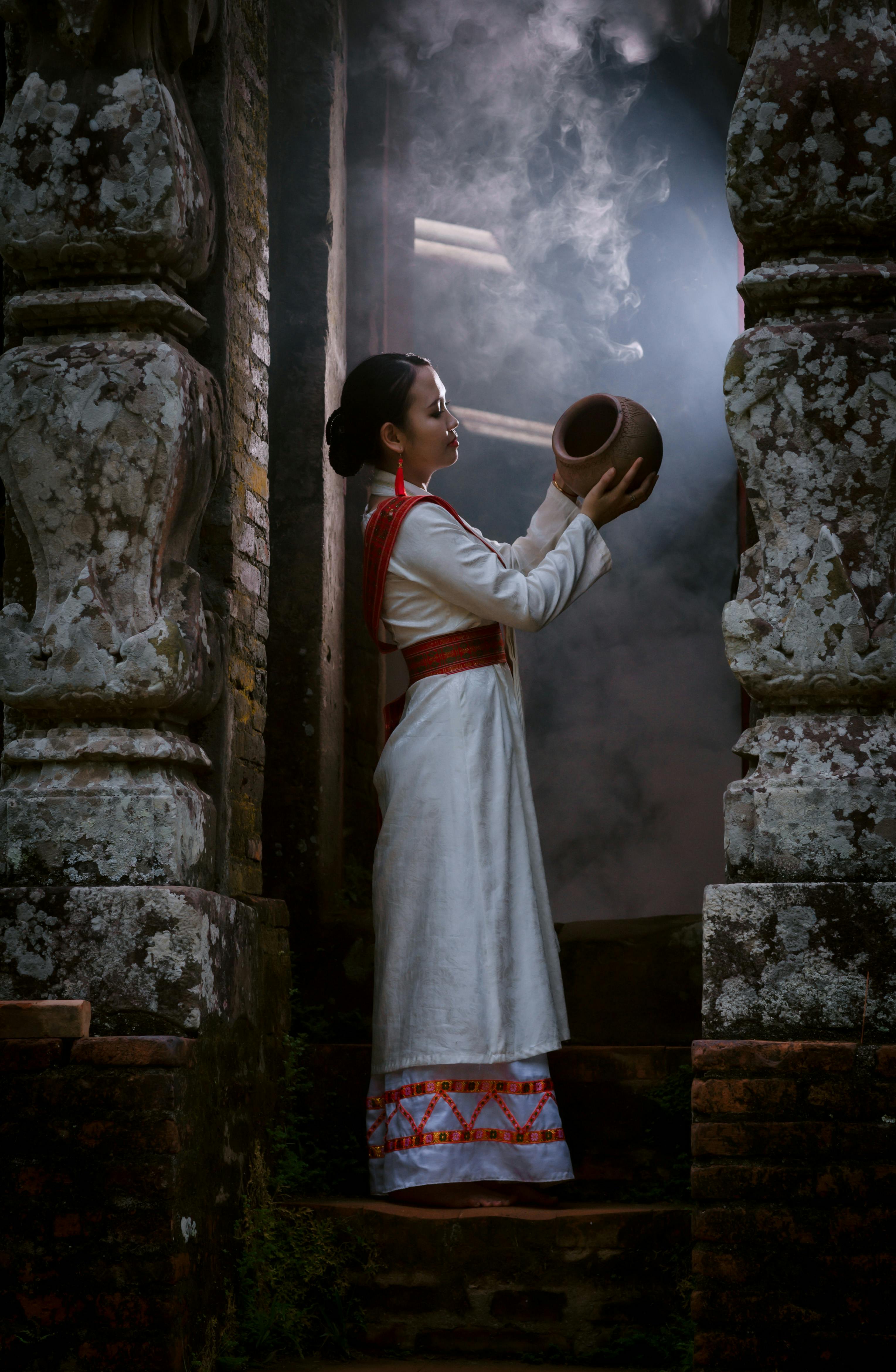 A woman in traditional attire holds a clay pot in an ancient temple setting, enveloped in mystical smoke.