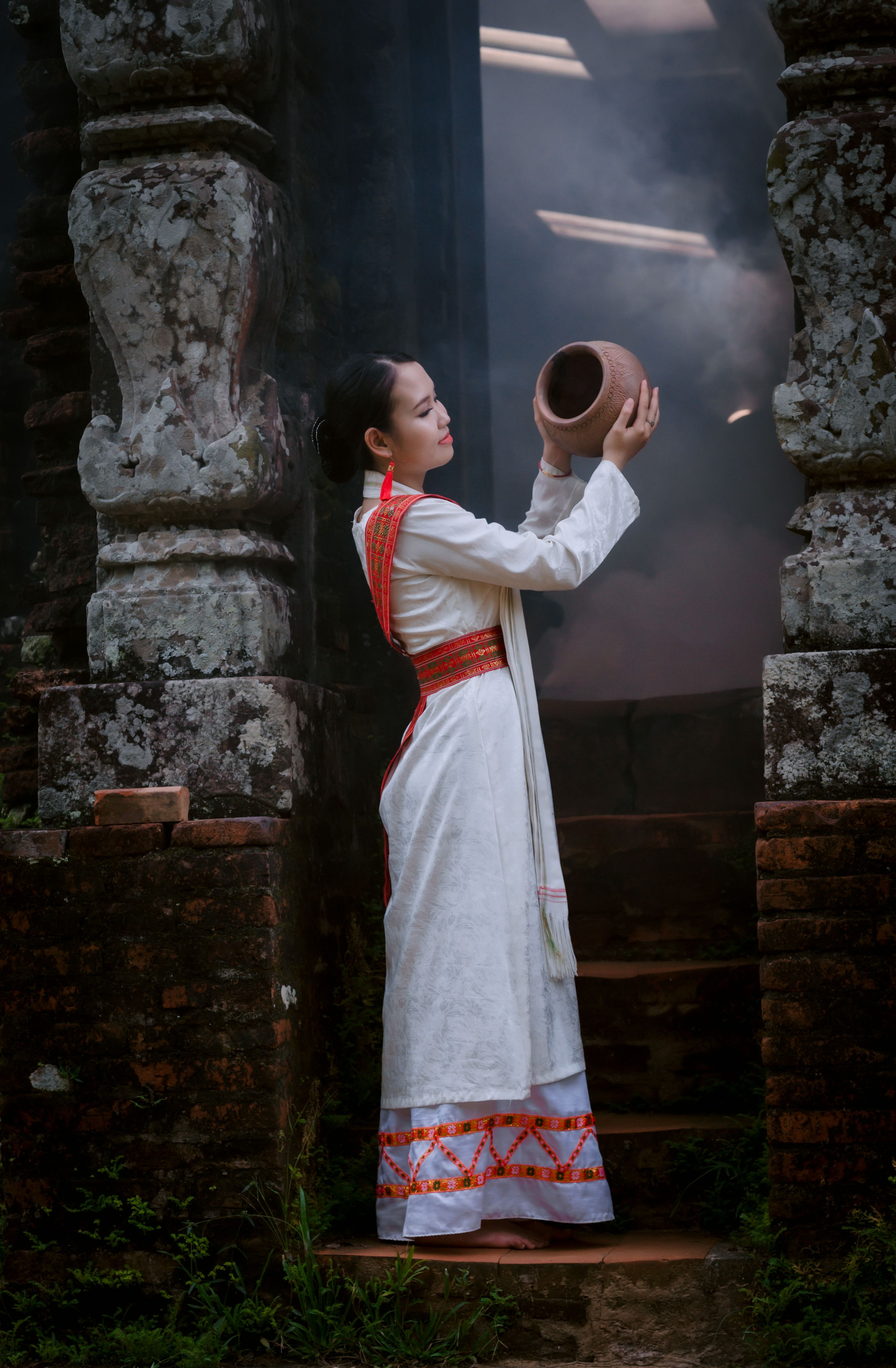 A woman in traditional attire holds a clay pot next to a weathered stone pillar, enveloped in mist.