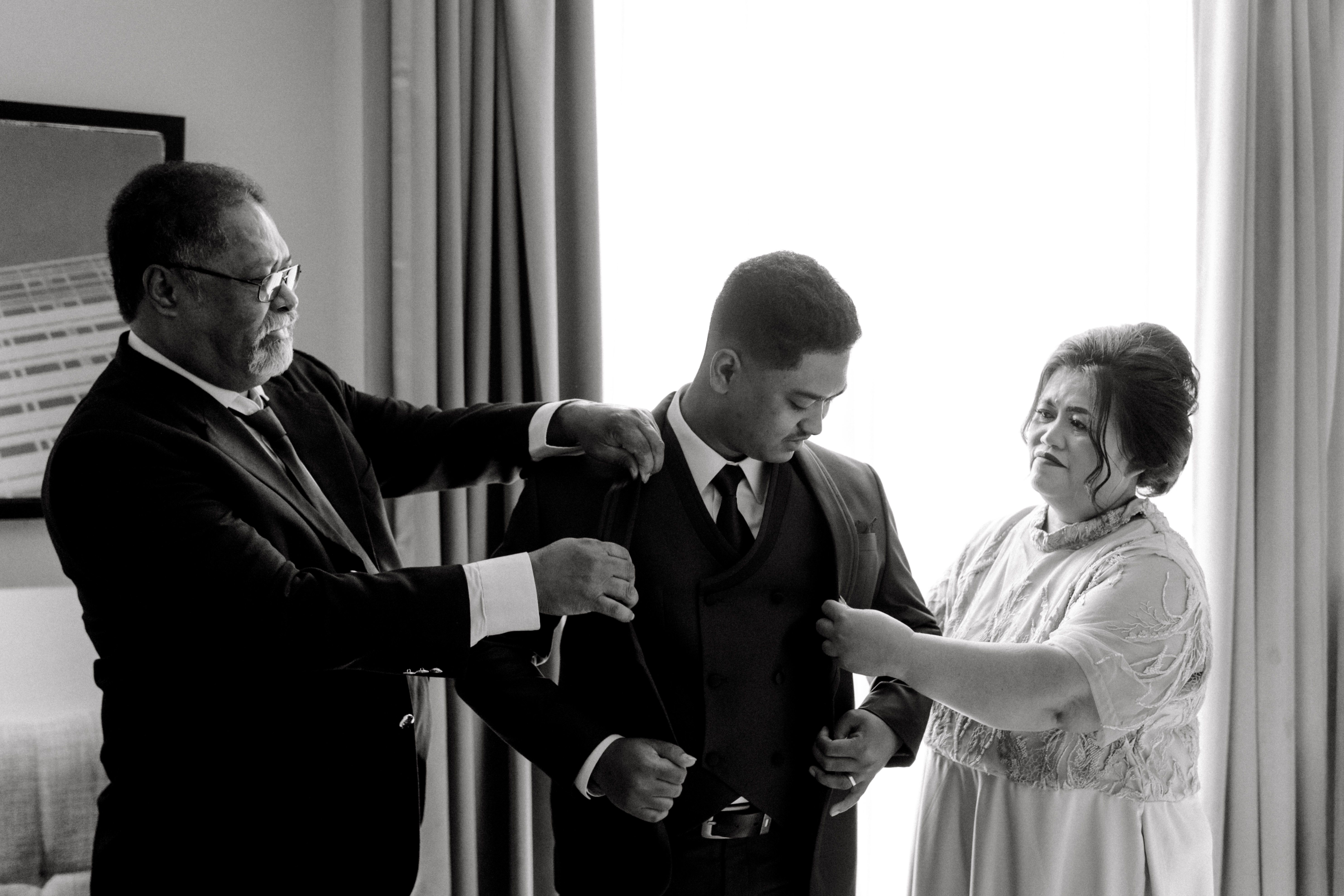 A black and white photo capturing a groom being assisted by family before the wedding ceremony.