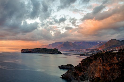 Dramatic coastal view with islands, mountains, and ocean at sunset under a cloudy sky.