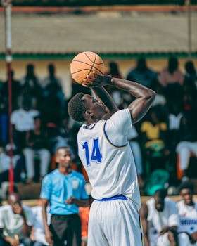A basketball player in white jersey number 14 shooting during a daytime game outdoors.
